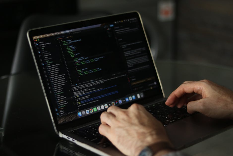 A close-up view of a person's hands working on a laptop, displaying a dark-themed coding interface with lines of colorful code, set on a dark desk surface indoors. The individual appears to be focused